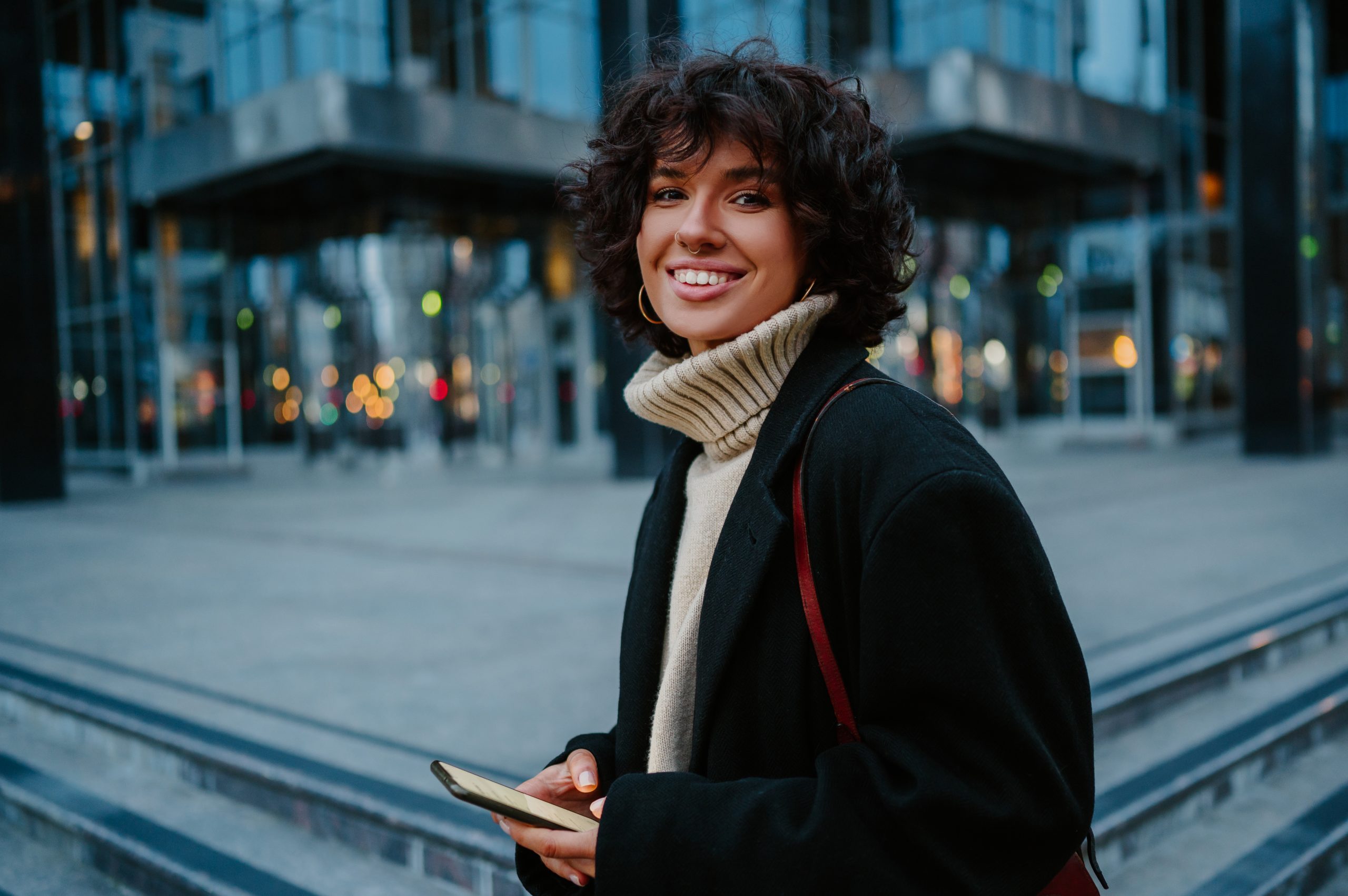 Portrait of a young dark haired curly girl looking straight to the camera with a big smile on her face while holding her smart phone in her hands and standing outside in front of a glass corporate building.