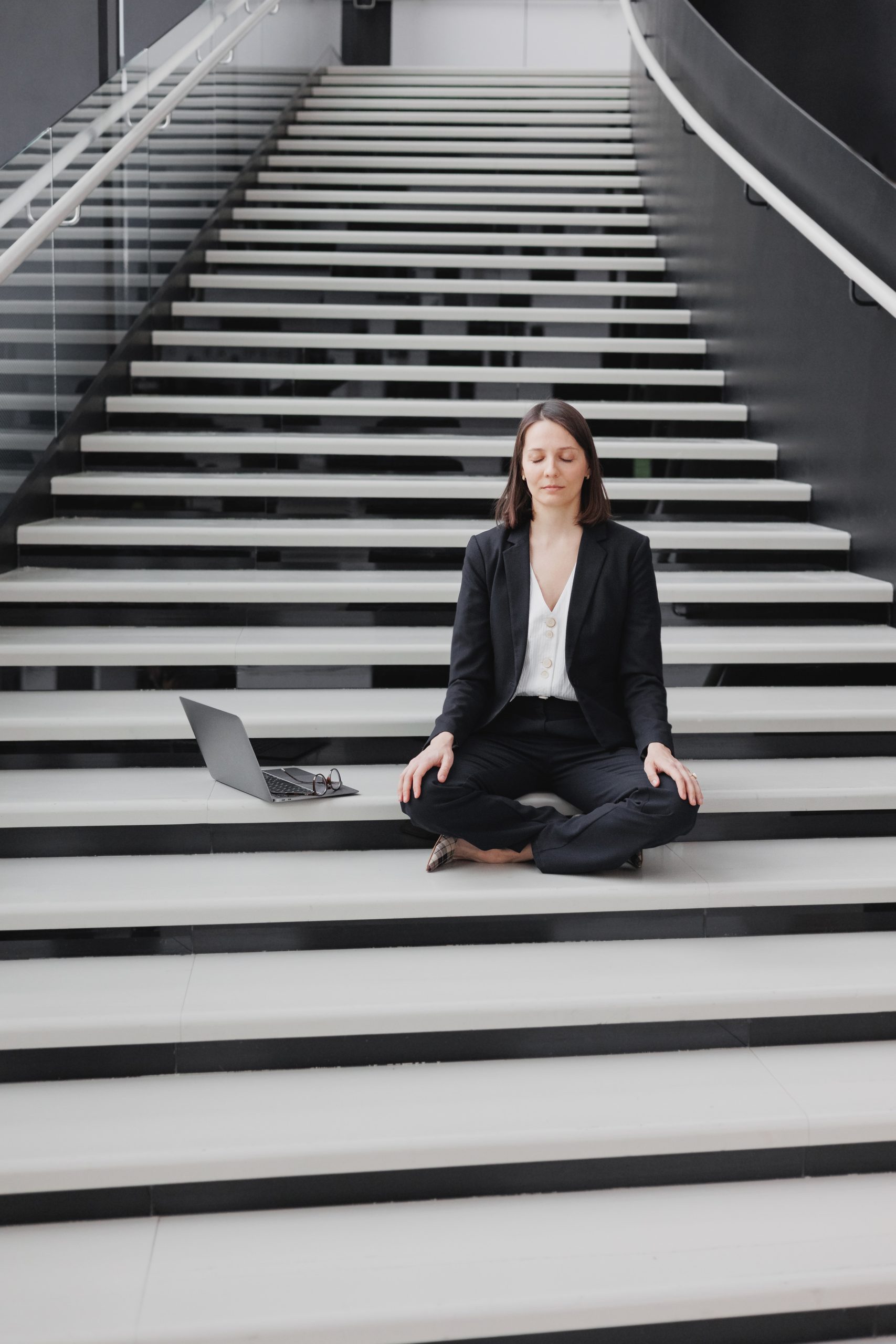 A woman dressed in business attire pauses for a moment of peace on the stairs of a workplace. Her laptop is placed carefully to her side while she takes a moment to close her eyes and break away from the screen and hyper-connectivity.