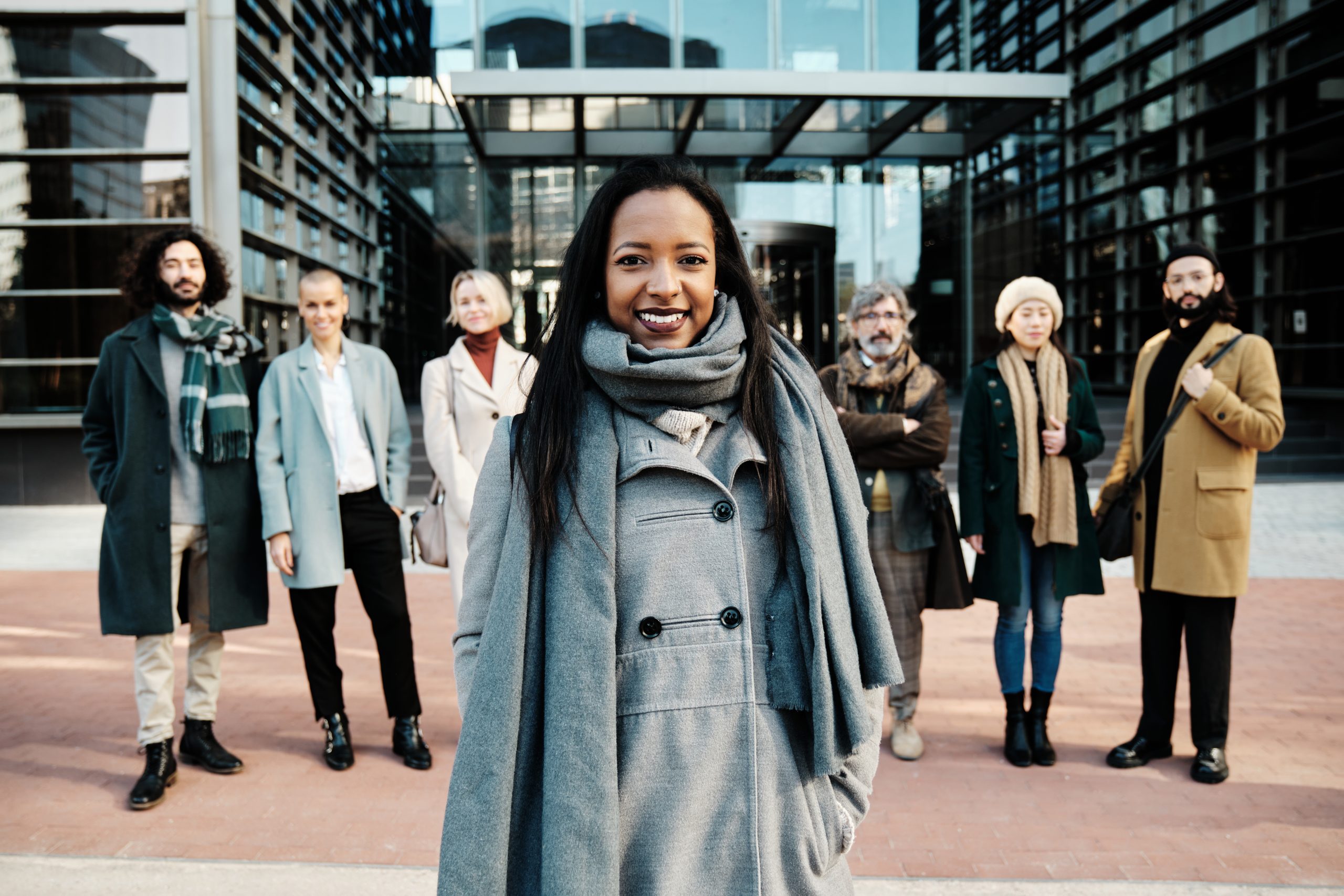 A woman of color in winter attire and corporate vibe stands in front of her team of six individuals. The six individuals are a mixture of men and women of varying ethnicities. The all stand confidently in front of a large glass corporate building.