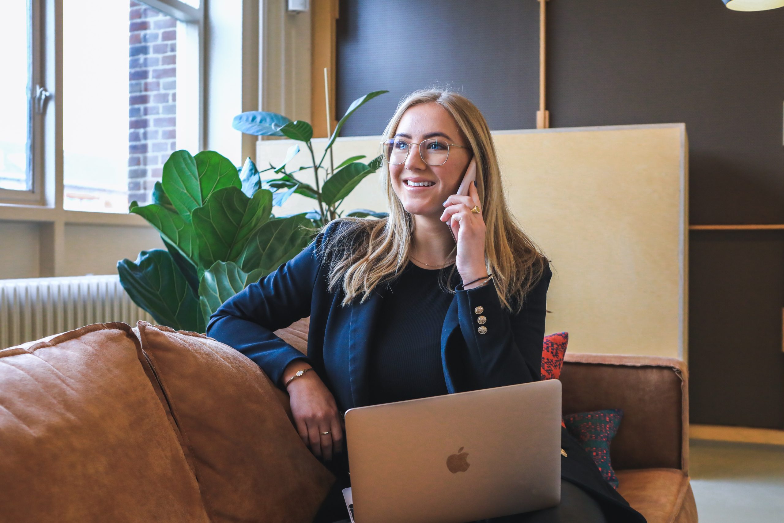 A blonde woman in corporate attire perches on a suede sofa while she takes a phone call on her mobile device. Her laptop is open and resting on her knees.