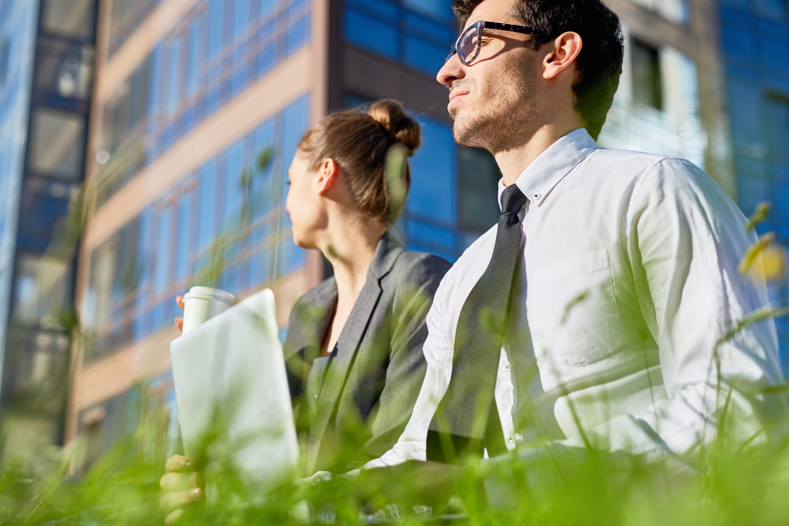 An executive man and executive woman sit outside with grass in view while they consider their professional topics. The woman holds a cup of coffee. The man is wearing a button-up and tie.