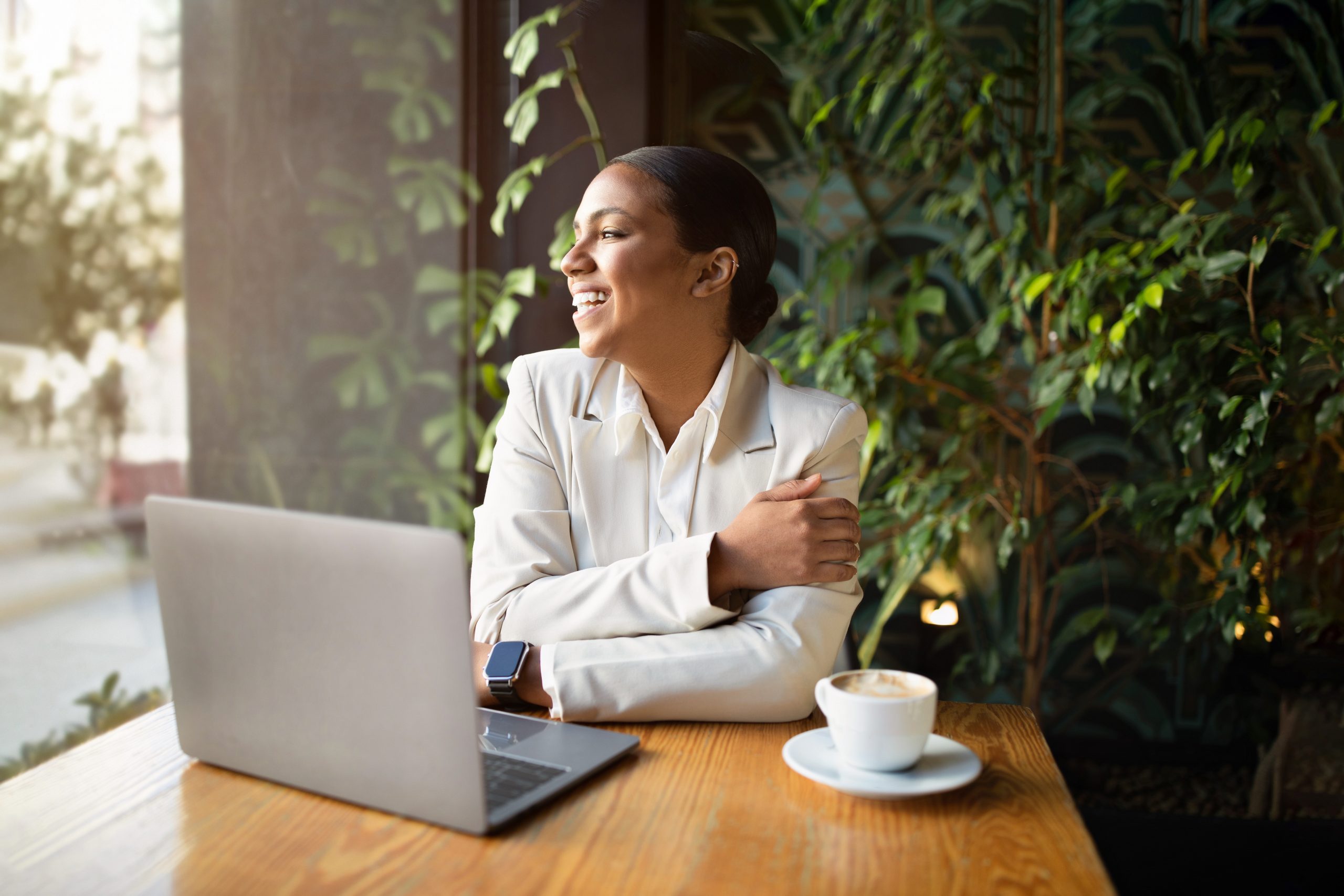 Smiling, young woman of color in suit with a smart watch works with laptop and enjoys cup of coffee