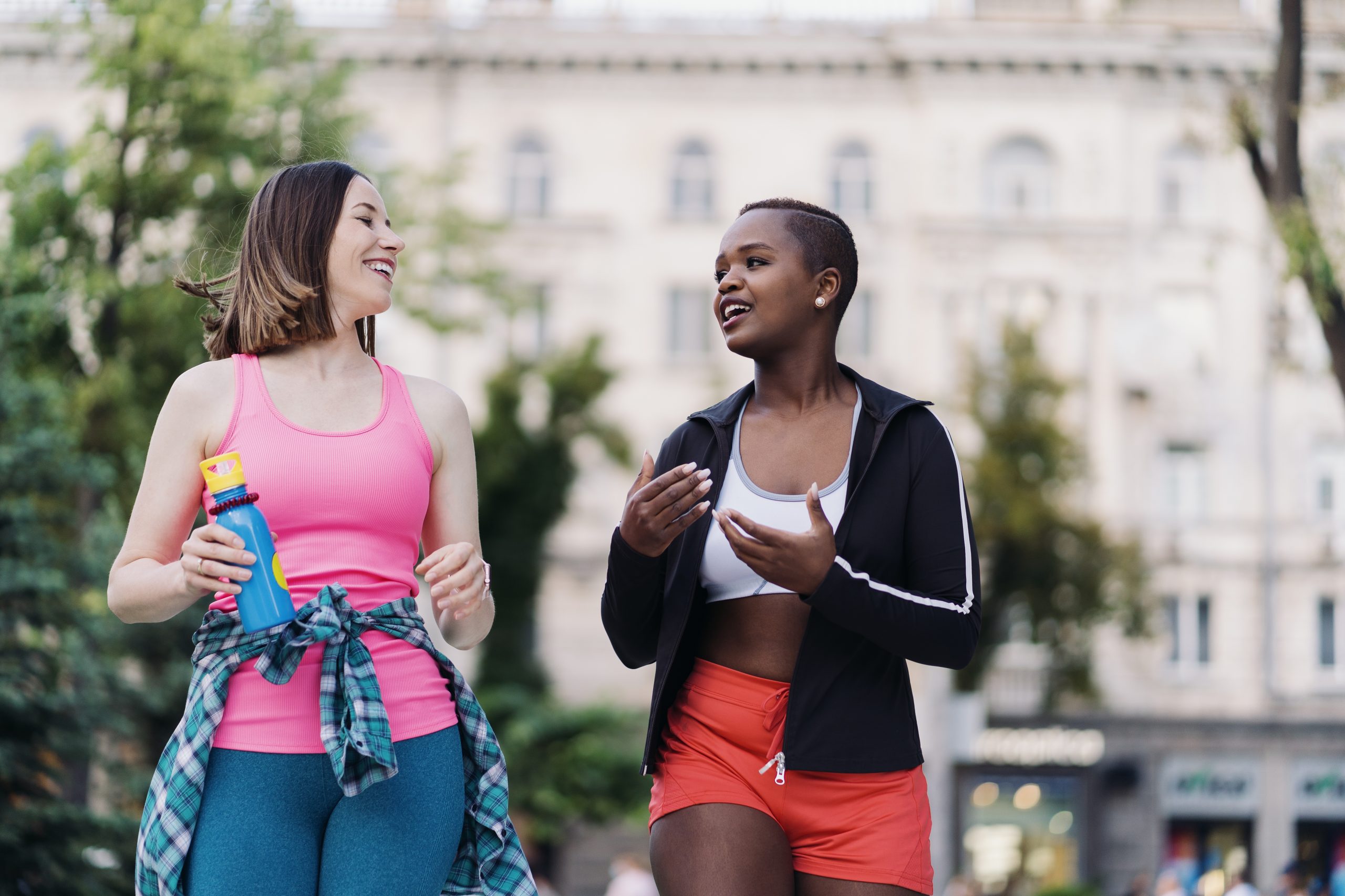 Two young, ethnically diverse women walk for exercise in fitness apparel while in active discussion with joy as the demeanor.