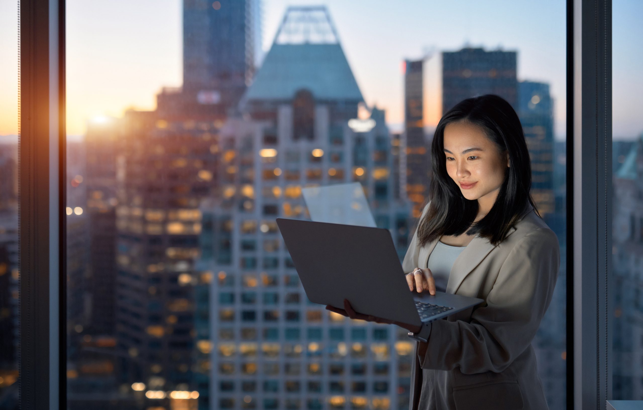 Young, busy Asian businesswoman executive working on laptop at night in dark corporate office. She is standing at a large window with big city evening view.